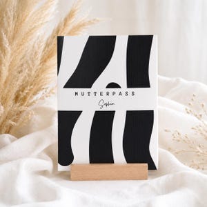 Könnte beinhalten: Eine rechteckige Karte mit einem schwarz-weißen Wellenstreifenmuster, die den Text MUTTERPASS und den Namen Sophia in einer Schreibschrift zeigt. Die Karte wird von einem kleinen, rechteckigen hellen Holzständer aufrecht gehalten.