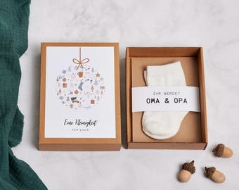 Puede incluir: Una caja de regalo con un calcetín de bebé blanco y una tarjeta. La tarjeta tiene un diseño de adorno navideño con el texto "Eine Kleinigkeit für Euch". El calcetín está en una caja marrón con la etiqueta "IHR WERDET OMA & OPA".