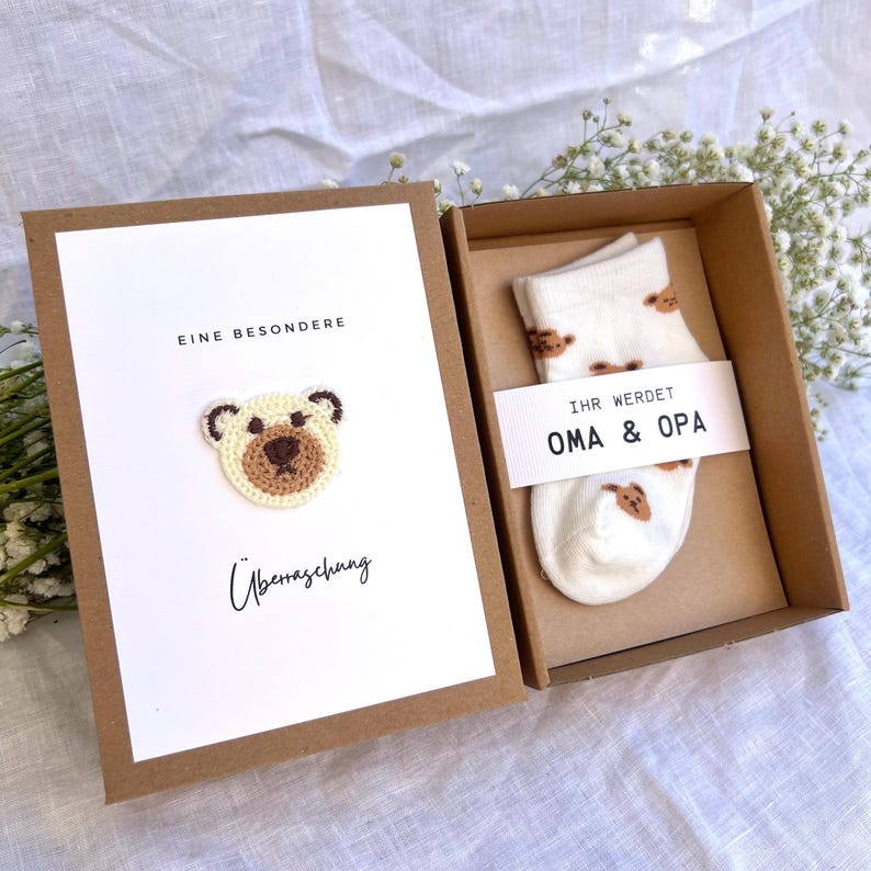May include: A gift box with a white card featuring a crocheted bear and the words "EINE BESONDERE &Uuml;berraschung". Inside, a pair of white socks with brown bear designs and a tag reading "IHR WERDET OMA & OPA".