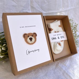 May include: A gift box with a white card featuring a crocheted teddy bear and the words "EINE BESONDERE &Uuml;berraschung". Inside, a pair of white socks with bear patterns and a tag that reads "IHR WERDET OMA & OPA".