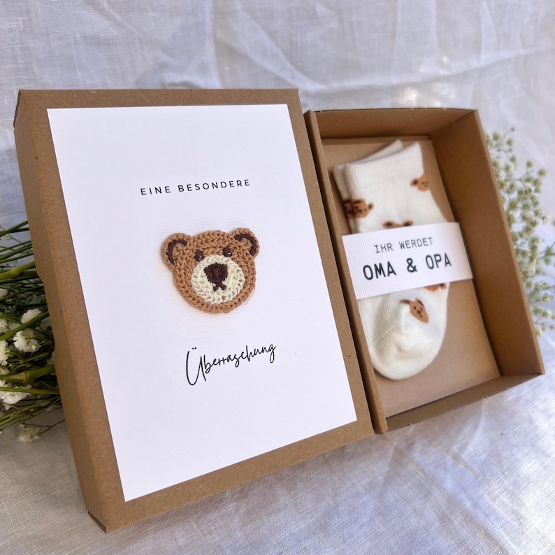May include: A gift box with a white card featuring a crocheted teddy bear and the words "EINE BESONDERE &Uuml;berraschung". Inside, a pair of white socks with bear patterns and a tag that reads "IHR WERDET OMA & OPA".