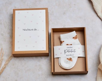 May include: A brown box with a white card that says "Möchtest du..." with pink hearts. A smaller brown box with a white sock with brown bear print and a white label that says "meine Patenante werden?" with pink hearts.