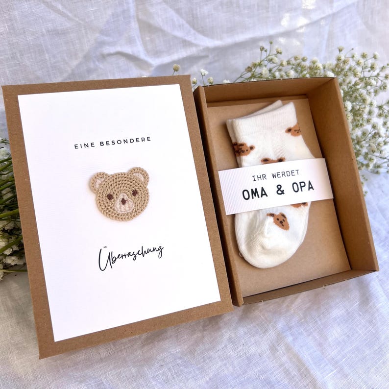 May include: A gift box with a greeting card and baby socks. The card has a crocheted teddy bear and the words "EINE BESONDERE &Uuml;berraschung". The socks are white with brown teddy bear patterns and the words "IHR WERDET OMA & OPA".