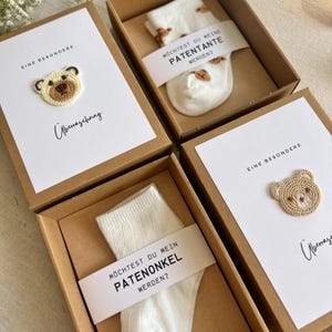 May include: Four gift boxes with white cards and socks. Each box has a brown cardboard exterior. The cards have a crocheted bear and the text "EINE BESONDERE" and "&Uuml;berraschung". The socks have a bear pattern and a banner that says "M&Ouml;CHTEST DU MEINE PATENTANTE WERDEN?" or "PATENONKEL WERDEN?"