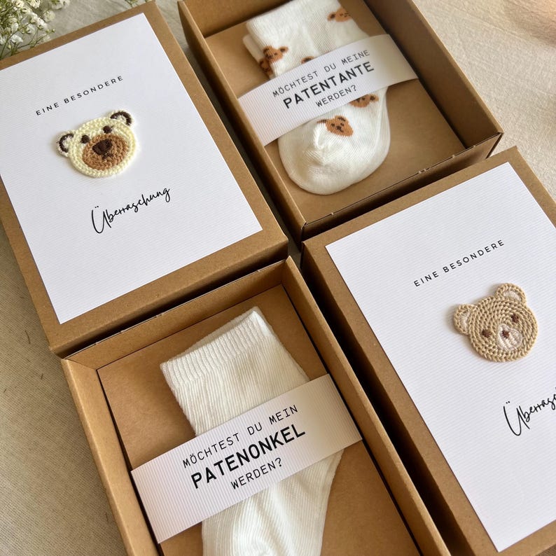 May include: Four gift boxes with white cards and socks. Each box has a brown cardboard exterior. The cards have a crocheted bear and the text "EINE BESONDERE" and "&Uuml;berraschung". The socks have a bear pattern and a banner that says "M&Ouml;CHTEST DU MEINE PATENTANTE WERDEN?" or "PATENONKEL WERDEN?"