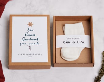 Puede incluir: Una caja de regalo con un calcetín de bebé blanco y una tarjeta. La tarjeta tiene texto en alemán, incluyendo "Ein kleiner Geschenk für euch" y un diseño de estrella. El calcetín está en una caja marrón, con el texto "IHR WERDET OMA & OPA" en una banda blanca.