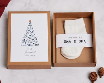Puede incluir: Una caja de regalo marrón con un calcetín blanco y una tarjeta. La tarjeta presenta un diseño de árbol de Navidad con hojas azules y verdes y el texto "Eine Kleinigkeit für Euch". El calcetín tiene una banda blanca con el texto "IHR WERDET OMA & OPA".