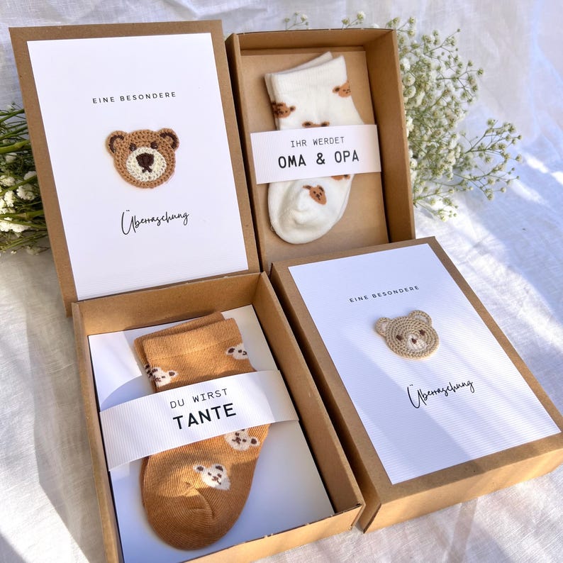 May include: Four gift boxes with cards and socks. The cards have a crocheted bear and text. One box contains white socks with bear designs and the text "IHR WERDET OMA & OPA". Another box has brown socks with bear designs and the text "DU WIRST TANTE".