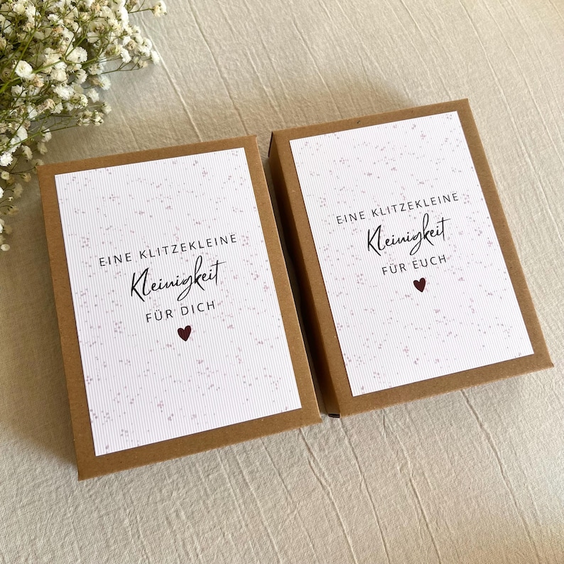 May include: Two rectangular brown cardboard gift boxes with white labels featuring a speckled pattern. The left label reads EINE KLITZEKLEINE Kleinigkeit F&Uuml;R DICH with a heart, and the right reads EINE KLITZEKLEINE Kleinigkeit F&Uuml;R EUCH with a heart.