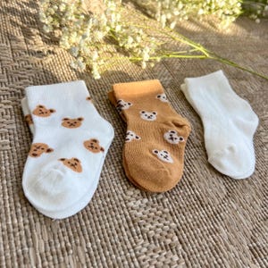 May include: Three pairs of socks: one white pair with a brown bear face pattern, one tan pair with a white bear face pattern, and one plain white pair. The socks feature a ribbed texture and are arranged side by side on a woven surface.