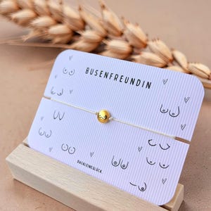 Könnte beinhalten: Ein zartes Armband mit einer einzelnen goldfarbenen Kugelperle, die eine kleine schwarze Strichzeichnung von Brüsten trägt, befestigt auf einer weißen Präsentationskarte. Die Karte ist mit BUSENFREUNDIN und DASKLEINEGLÜCK sowie kleinen Herz- und Brustmotiven bedruckt.