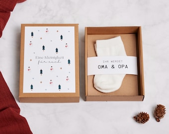 Puede incluir: Una caja de regalo con un calcetín blanco en su interior, presentada en una caja de cartón marrón. La caja tiene una tarjeta decorativa con un patrón de árboles de Navidad y muñecos de nieve. El texto en la tarjeta dice "Eine Kleinigkeit für euch". El calcetín tiene una banda blanca con el texto "IHR WERDET OMA & OPA".