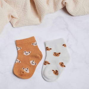 May include: Two pairs of baby socks. One pair is orange with white bear faces, and the other is white with brown bear faces. The socks are made of a soft material and are designed for comfort. The background includes a cream-colored fabric.