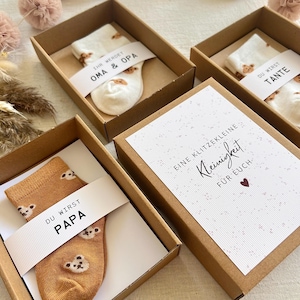 May include: Three brown cardboard gift boxes containing pairs of socks with bear patterns. Each box features a white paper band with text: 'IHR WERDET OMA & OPA', 'DU WIRST TANTE', and 'DU WIRST PAPA'. One box lid displays 'EINE KLITZEKLEINE Kleinigkeit F&Uuml;R EUCH'.