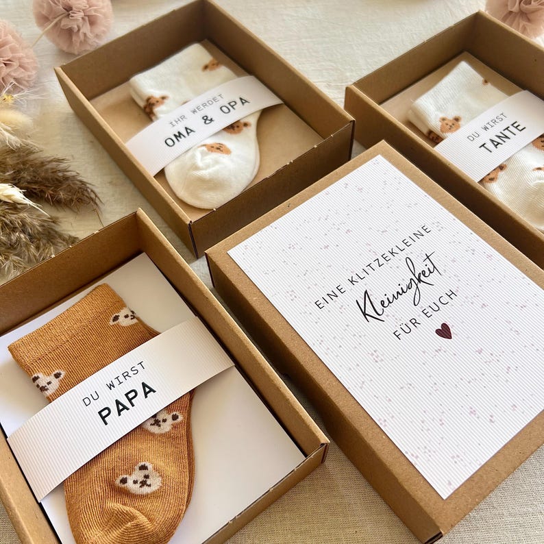 May include: Three brown cardboard gift boxes containing pairs of socks with bear patterns. Each box features a white paper band with text: 'IHR WERDET OMA & OPA', 'DU WIRST TANTE', and 'DU WIRST PAPA'. One box lid displays 'EINE KLITZEKLEINE Kleinigkeit F&Uuml;R EUCH'.