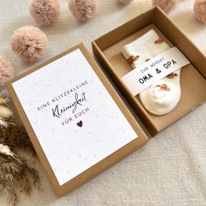 May include: A brown cardboard gift box containing a pair of white baby socks with a brown bear pattern. A paper band around the socks reads "IHR WERDET OMA & OPA". A card in the box lid reads "EINE KLITZEKLEINE Kleinigkeit F&Uuml;R EUCH" with a heart.