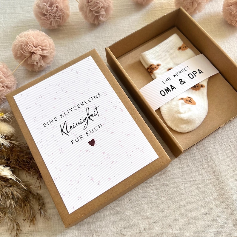 May include: A brown cardboard gift box containing a pair of white baby socks with a brown bear pattern. A paper band around the socks reads "IHR WERDET OMA & OPA". A card in the box lid reads "EINE KLITZEKLEINE Kleinigkeit F&Uuml;R EUCH" with a heart.
