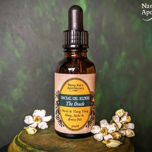 May include: A brown glass bottle with a dropper top, labelled "Facial Oil Elixir The Oracle" with a list of ingredients including Neroli & Ylang Ylang, Hemp, Jojoba & Arnica Oils. The bottle is sitting on a wooden surface with white flowers.