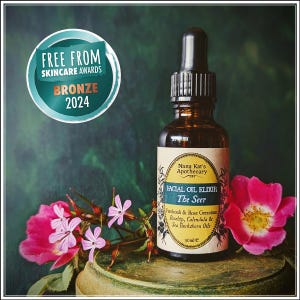 May include: A brown glass bottle with a dropper top, labelled "Nana Kat's Apothecary Facial Oil Elixir The Seer Patchouli & Rose Geranium Rosehip, Calendula & Sea Buckthorn Oils 30ml e". The bottle is sitting on a wooden surface with pink flowers.