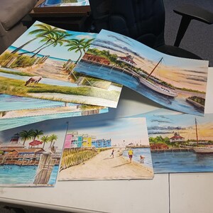 May include: A stack of colorful paintings depicting various scenes of a lighthouse, a boat, and a beach. The paintings are on a white table with a white chair in the background.