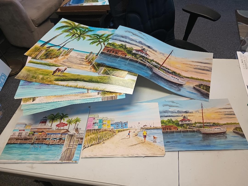 May include: A stack of colorful paintings depicting various scenes of a lighthouse, a boat, and a beach. The paintings are on a white table with a white chair in the background.