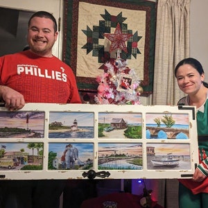 May include: A couple poses with a framed artwork featuring eight watercolor paintings of coastal scenes. The artwork is framed in a white, distressed wood frame with a black border. The couple is standing in a room decorated for Christmas with a small Christmas tree and a wreath.
