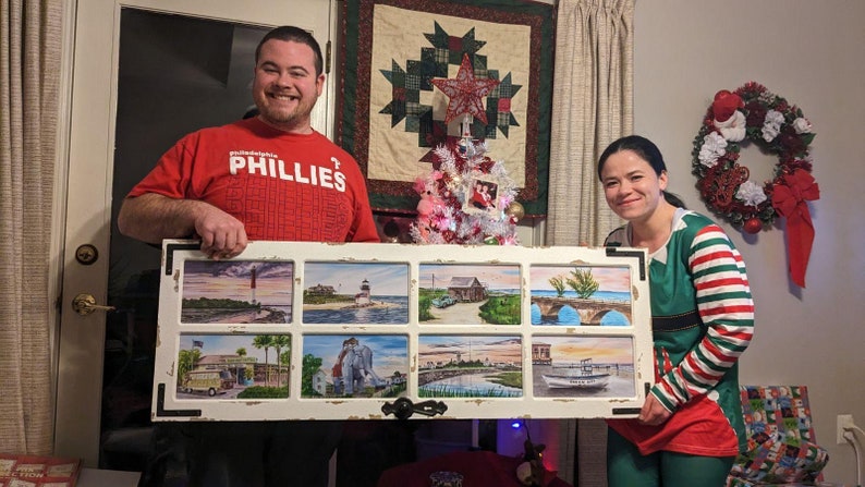 May include: A couple poses with a framed artwork featuring eight watercolor paintings of coastal scenes. The artwork is framed in a white, distressed wood frame with a black border. The couple is standing in a room decorated for Christmas with a small Christmas tree and a wreath.
