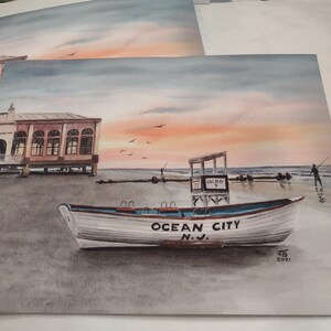 May include: Watercolor painting of a white boat with "Ocean City N.J." written on the side, sitting on a sandy beach with a lifeguard stand and a building in the background. The sky is a soft pink and blue.