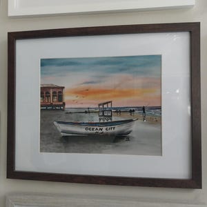 May include: A watercolor painting of a white boat with the words "Ocean City, Md." on the side. The boat is on a beach with a building in the background and a sunset in the sky.