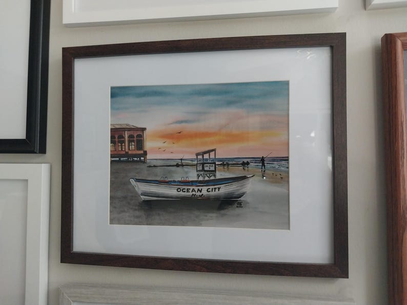 May include: A watercolor painting of a white boat with the words "Ocean City, Md." on the side. The boat is on a beach with a building in the background and a sunset in the sky.
