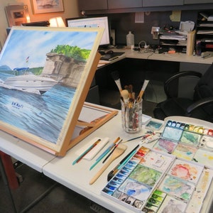 May include: A watercolor painting of a boat on a lake with a cliff in the background. The painting is on an easel and there are watercolor paints and brushes on a table next to it.