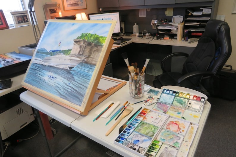 May include: A watercolor painting of a boat on a lake with a cliff in the background. The painting is on an easel and there are watercolor paints and brushes on a table next to it.