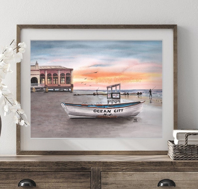 May include: Watercolor painting of a boat on a beach at sunset. The boat is white with blue lettering that says "Ocean City, N.J." The painting is framed in a brown frame.
