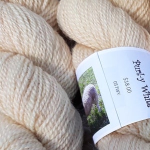 May include: Close-up of ivory-colored yarn skeins. A label reads "Purly White" with a picture of a sheep. The yarn is thick and appears soft, ideal for knitting or crocheting projects. The label also includes the number "057WY".