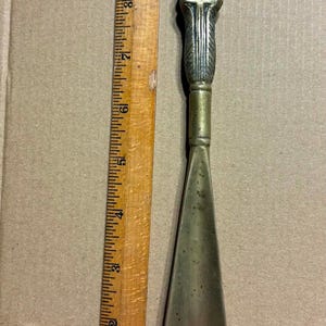 May include: A brass shoehorn featuring a sculpted horse head handle, positioned next to a wooden ruler showing a total length of approximately 11 inches. The metal surface displays a metallic gold tone with visible texture and aged patina.