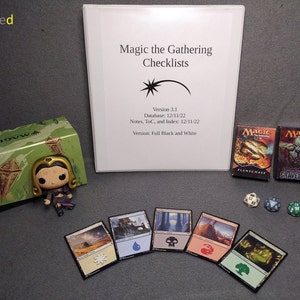 May include: A green and brown box with the word "Magic" printed on it. A small figurine of a woman with a gold crown and purple clothing. A white sheet of paper with the text "Magic the Gathering Checklists" printed on it. The text "Version 3.1" is printed below the title. The text "Database: 12/11/22" is printed below the version number. The text "Notes, ToC, and Index: 12/11/22" is printed below the database date. The text "Version: Full Black and White" is printed below the notes date. Two Magic the Gathering cards are visible. The card on the left is titled "Flamewake" and the card on the right is titled "Graveborn". There are three dice on the table. One is red, one is blue, and one is green.