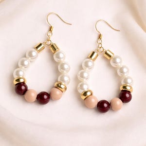 May include: A pair of teardrop-shaped earrings. Each earring features a gold-colored hook and a design of white, blush, and burgundy-colored beads. Gold-colored metal accents separate the bead colors. The earrings are displayed on a white fabric.