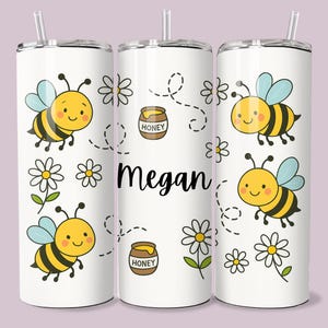 May include: White tumbler with a bee-themed design. The tumbler features cartoon bees, daisies, and honey pots. The name "Megan" is printed in black. Includes a straw.