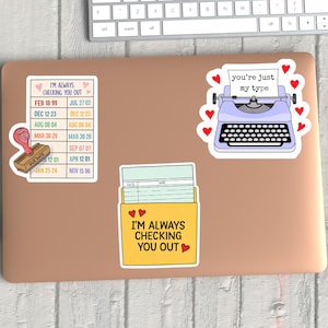 May include: Laptop covered with stickers. One sticker says "I'M ALWAYS CHECKING YOU OUT" with a list of dates and a stamp. Another sticker is a light purple typewriter with the text "you're just my type". A third sticker says "I'M ALWAYS CHECKING YOU OUT".