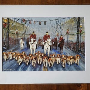 May include: A framed print depicts a winter scene with riders on horseback, accompanied by a pack of hounds. The riders wear red coats, and the dogs are brown and white. The scene is set on a snowy street with decorated lampposts.