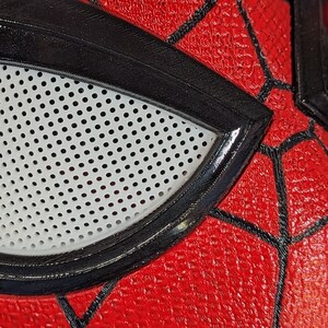 Puede incluir: Primer plano de una m&aacute;scara de Spider-Man roja y negra con una abertura para los ojos de malla blanca.