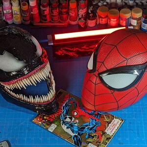 Puede incluir: Una m&aacute;scara negra de Venom con dientes afilados y una m&aacute;scara roja y negra de Spider-Man. Las m&aacute;scaras est&aacute;n sobre una superficie azul con un c&oacute;mic que presenta a Spider-Man y Venom.