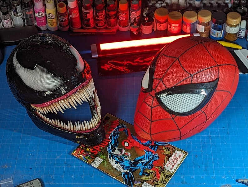 Puede incluir: Una m&aacute;scara negra de Venom con dientes afilados y una m&aacute;scara roja y negra de Spider-Man. Las m&aacute;scaras est&aacute;n sobre una superficie azul con un c&oacute;mic que presenta a Spider-Man y Venom.