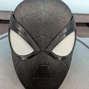 Puede incluir: M&aacute;scara de Spider-Man en blanco y negro con una superficie texturizada. La m&aacute;scara tiene un &aacute;rea de ojos blanca con un contorno negro y un dise&ntilde;o de telara&ntilde;a negra.