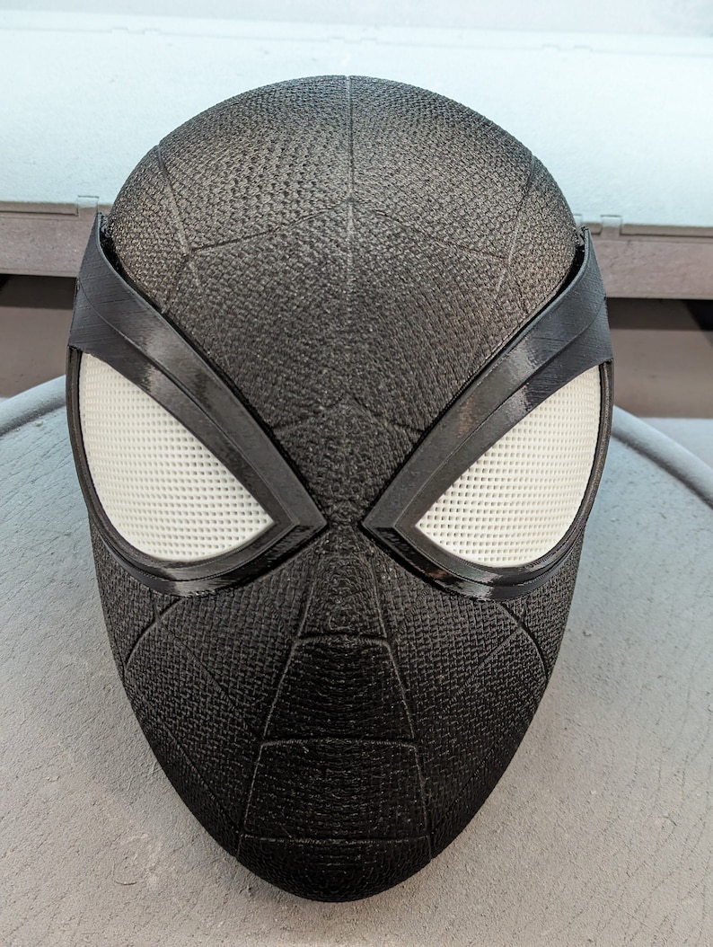 Puede incluir: M&aacute;scara de Spider-Man en blanco y negro con una superficie texturizada. La m&aacute;scara tiene un &aacute;rea de ojos blanca con un contorno negro y un dise&ntilde;o de telara&ntilde;a negra.