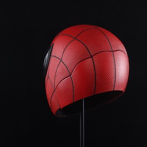 Puede incluir: Un casco rojo y negro de Spider-Man con una superficie texturizada. El casco est&aacute; en un soporte negro.