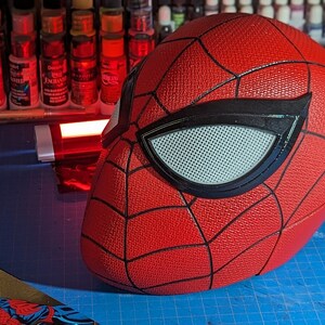 Puede incluir: Una m&aacute;scara de Spider-Man roja y negra con una superficie texturizada y una pieza de ojo de malla blanca.