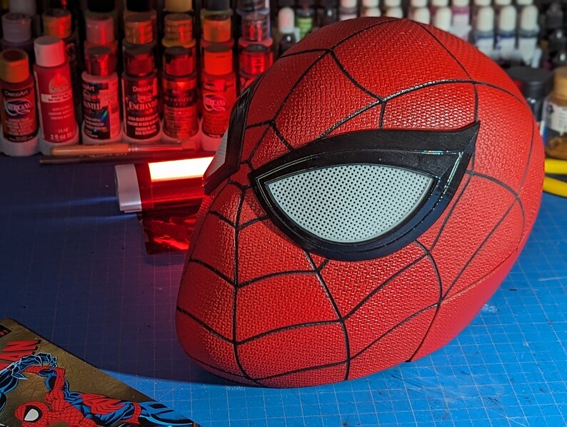 Puede incluir: Una m&aacute;scara de Spider-Man roja y negra con una superficie texturizada y una pieza de ojo de malla blanca.