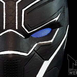 K&ouml;nnte beinhalten: Nahaufnahme einer schwarzen und wei&szlig;en Plastikmaske von Black Panther mit blauen LED-Augen. Die Maske hat eine strukturierte Oberfl&auml;che und einen wei&szlig;en Umriss um die Augen.