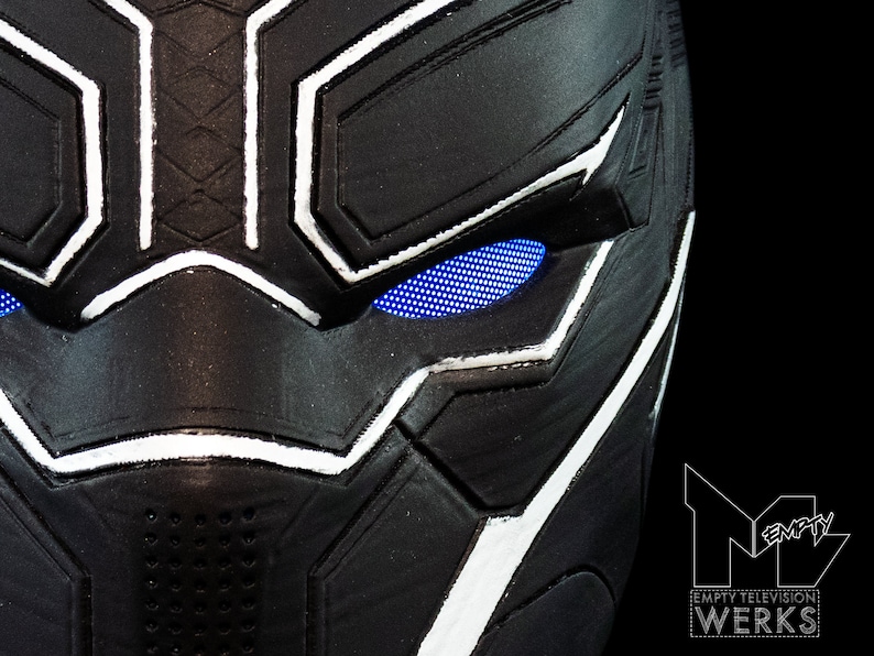 K&ouml;nnte beinhalten: Nahaufnahme einer schwarzen und wei&szlig;en Plastikmaske von Black Panther mit blauen LED-Augen. Die Maske hat eine strukturierte Oberfl&auml;che und einen wei&szlig;en Umriss um die Augen.